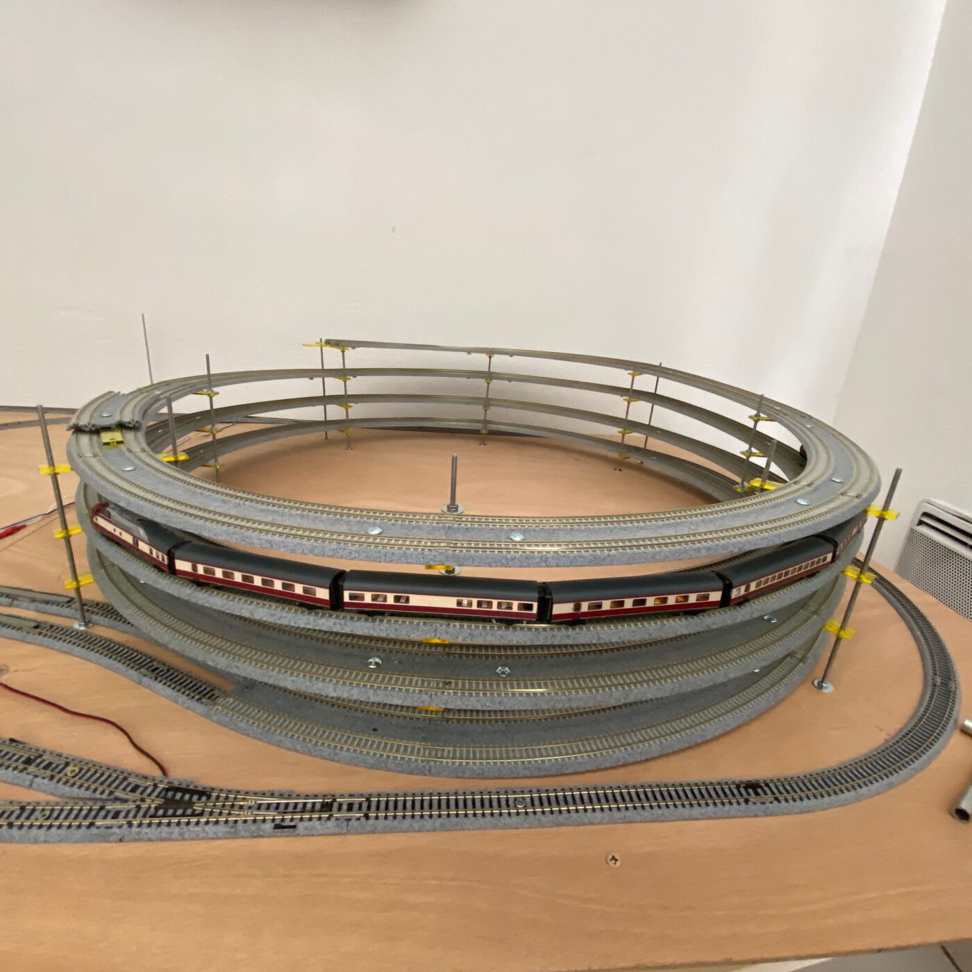 Hélicoïde de réseau ferroviaire miniature à plusieurs niveaux, posé sur une table en contreplaqué, avec un train de voitures voyageurs rouge et crème qui circule sur le dernier anneau.