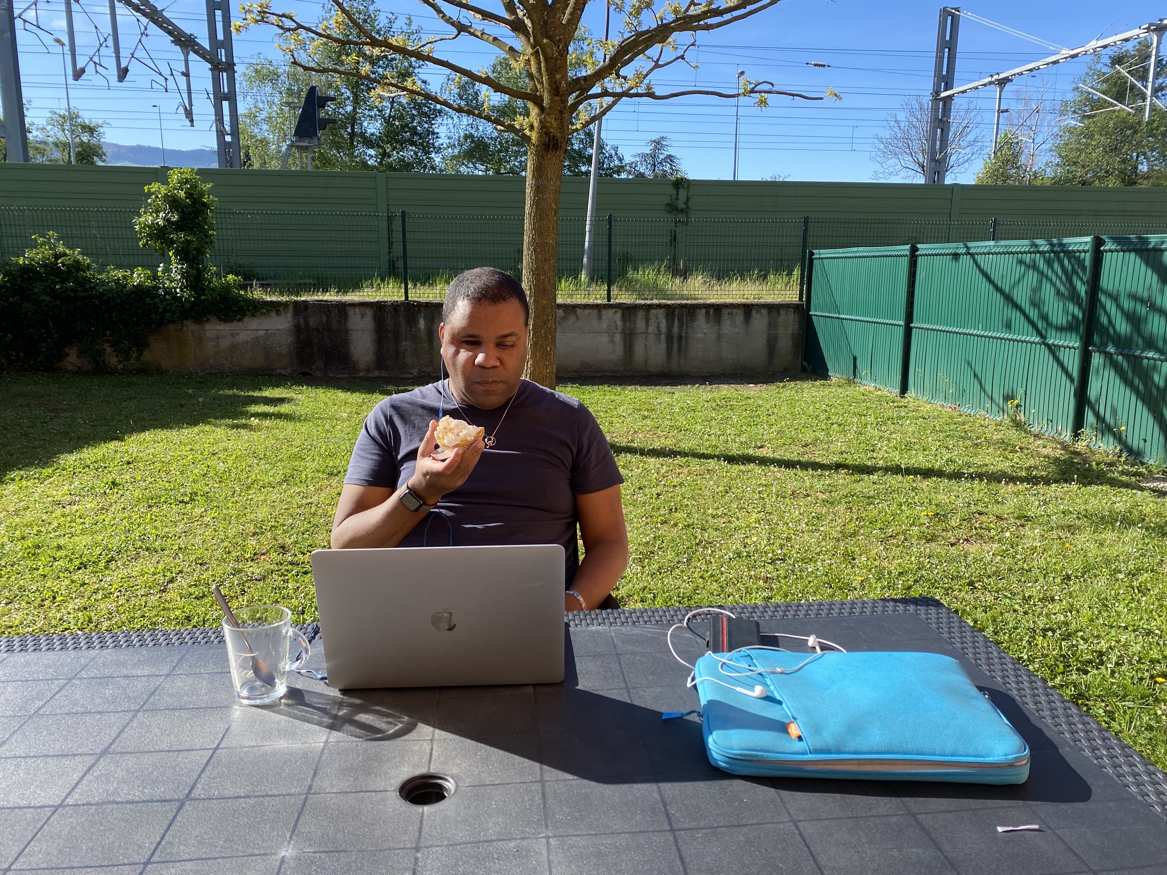Un homme est assis à une table de jardin, travaillant sur un ordinateur portable tout en tenant un morceau de gateau. Sur la table se trouvent un verre avec une cuillère et une housse d’ordinateur bleue. La scène se déroule dans un jardin ensoleillé avec un arbre derrière lui et une ligne ferroviaire visible en arrière plan.