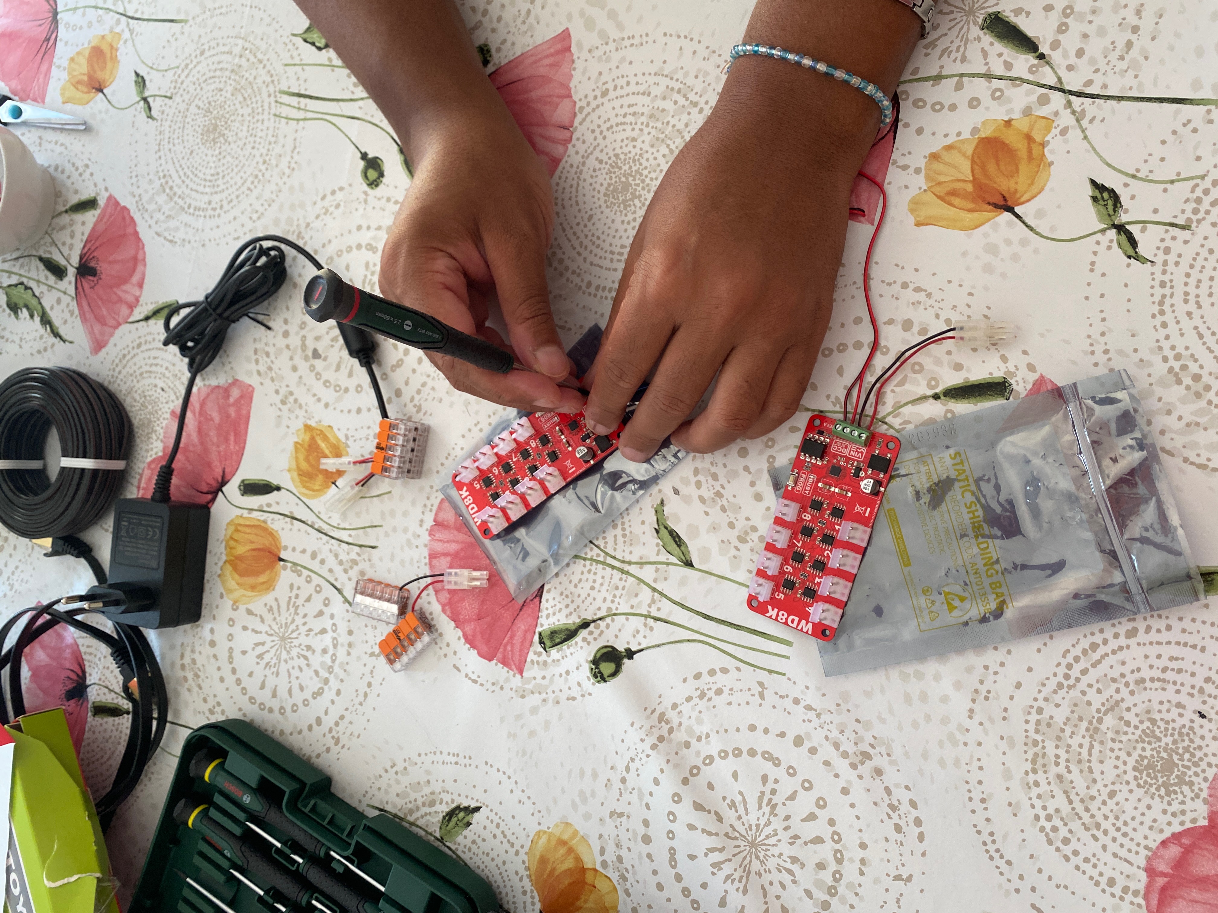 sur une tabl, quelqu'un travaille sur un circuit imprimé rouge avec un tournevis de précision. Autour, coffret de tournevis, bobine de câble, alimentation, connecteurs rapides et sachet antistatique.